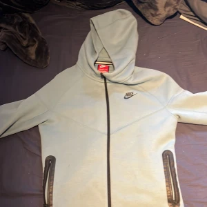 Ljusgrå Nike hoodie med dragkedja XL - Nike hoodie i ljusgrått med svart dragkedja och huva. Tröjan har två fickor med dragkedja framtill och klassisk Nike-logga på bröstet. 