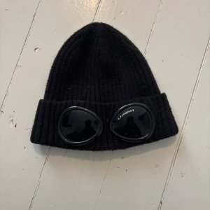 Svart ribbstickad mössa C.P. Company - Svart ribbstickad mössa från C.P. Company med ikoniska goggles framtill på uppviket. Tillverkad i mjuk ull och har en one size passform.Perfekt accessoar. 9/10 skick!
