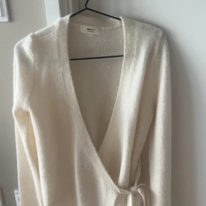 Vit omlottkofta från Gina Tricot - Supermjuk vit omlottkofta från Gina Tricot i ullblandning. Koftan har långa ärmar, knytband i midjan och en djup v-ringning. Perfekt för lager-på-lager och chill höst- eller vinterstil.