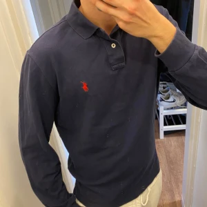 Mörkblå långärmad Pikétröja från Ralph Lauren - Klassisk mörkblå långärmad piké från Polo Ralph Lauren med röd broderad logga på bröstet. Tröjan har krage, knappar vid halsen och är tillverkad i bomull. Perfekt för en stilren och avslappnad look. Storlek L men passar mer som M. Snabb Leverans!