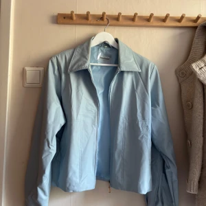 Ljusblå vindjacka från Neu View - Snygg ljusblå vindjacka från Neu View med klassisk krage och dragkedja framtill. Jackan har en clean look och är tillverkad i ett lätt syntetmaterial, perfekt för vår och sommar. Enkel och stilren design som passar till det mesta.