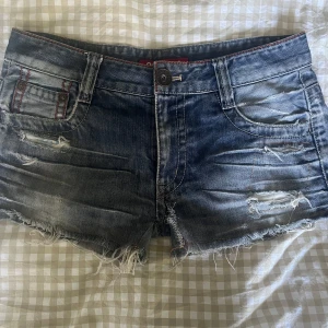 Blå slitna jeansshorts  - Säljer ett par blå jeansshorts med  fransiga kanter och slitna detaljer. Shortsen har klassiska fem fickor, röda sömmar och en cool tvättad look. Hål i dom som blivit lagat men gått upp. Lätt att laga skulle jag tro. Vet inte exakt storlek men jag har S och den är för stor för mig. 