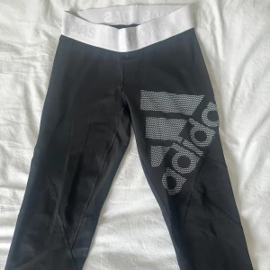 Svarta och  vita Adidas träningsbyxor - Snygga svarta leggings från Adidas med bred vit midja där Adidas-loggan är präglad. Ena benet har en stor vit Adidas-logga. Stretchigt material som sitter tight och är perfekt för träning eller chill. Supersnygga till sneakers
