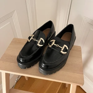 Svarta chunky loafers med gulddetalj strl 38 - Snygga svarta loafers med chunky sula och guldfärgad metallspänne framtill. Klassisk rund tå och robust platt sula ger en cool och modern look. Perfekta att styla till både jeans och kostymbyxor.
