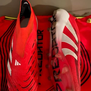 Adidas Predator Elit fotbollsskor - Säljer ett par Adidas Predator fotbollsskor i rött med vita detaljer och svarta inslag. Skorna har en strumpliknande passform utan snörning, mönstrad ovandel och tydliga dobbar för bästa grepp på planen. Perfekta för dig som vill sticka ut på matchen.