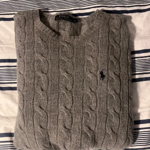 Grå kabelstickad tröja Polo Ralph Lauren - Kabelstickad grå tröja från Polo Ralph Lauren med klassisk rund halsringning och broderad marinblå logga på bröstet. Tröjan är i mjuk ullblandning och har långa ärmar samt ribbade muddar och nederkant. Perfekt för lager-på-lager under kyliga dagar. Bara att höra av er för frågor . Mvh Elton