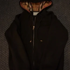 Svart Burberry hoodie med dragkedja - Svart hoodie från Burberry med klassiskt rutigt foder i huvan och broderad logga på bröstet. Tröjan har dragkedja framtill, snörning i huvan och guldfärgade detaljer på snörena. Perfekt för dig som vill ha en stilren och lyxig look.pris kan diskuteras!