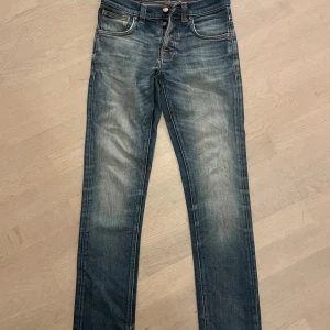 Blå jeans från Nudie Jeans - Säljer ett par klassiska blå jeans från Nudie Jeans med snygg slitning och kontrastsömmar. Modellen har raka ben, fem fickor och orange detaljer. Bakfickorna har Nudie Jeans karaktäristiska broderi. Perfekta för en avslappnad och trendig look. Från början är jeansen W28 L32. Men sedan så förkortade jag jeansen till ca L29