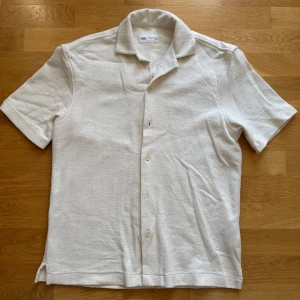 Beige kortärmad skjorta från Zara - Säljer en stilren beige skjorta från Zara i storlek S. Skjortan har korta ärmar, klassisk krage och knäppning framtill. Materialet är strukturerat och känns mjukt mot huden, perfekt för varmare dagar. Enkel och clean look som passar till det mesta.