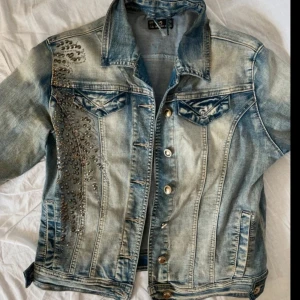 Jeansjacka med nitar från Diablo.z - Snygg ljusblå jeansjacka från Diabloz med coola nitar och stenar i mönster både fram och bak. Klassisk modell med knappar, bröstfickor och krage. Perfekt för dig som vill sticka ut med en unik detalj på jackan.