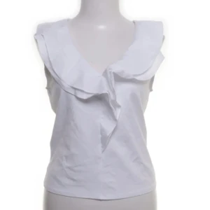 Vit volangblus från MNG XS - Supersöt vit blus från MNG i storlek XS. Ärmlös modell med stor volangkrage som ger en extra touch. Perfekt för dig som gillar en clean och chic look. Tillverkad i mjuk bomull som känns skön mot huden.LÅNAD BILD 