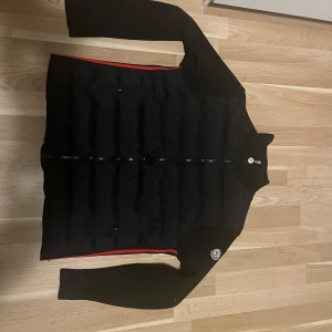 Svart Moncler pufferjacka med stickade ärmar - Svart pufferjacka från Moncler med stickade ärmar och klassisk Moncler-logga på ena ärmen. Jackan har dragkedja framtill och en röd detalj längs nederkanten. Perfekt för vintern och höst.pris går att diskutera.