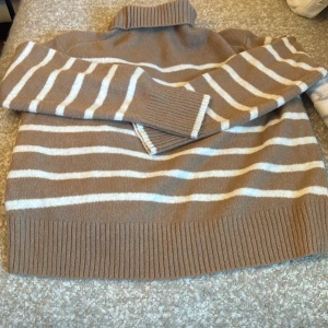 Randig polotröja i beige från Uniqlo - Supermysig polotröja från Uniqlo i beige med vita ränder. Tröjan har ribbade muddar och hög krage, perfekt för kyliga dagar. Den är stickad och har lång ärm, vilket gör den extra skön och stilren.