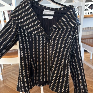 Randig svart och beige jacka från Zara - Säljer en snygg jacka från Zara med svart bas och beigea ränder. Jackan har bred krage, knappar framtill och ett bälte i midjan. Materialet är grovt och känns lite ulligt, perfekt för att ge en cool och trendig look. Jackan är rätt så liten i storleken, jag skulle säga att de är en ungefär M