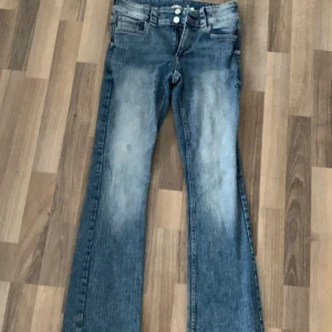 Blå bootcut jeans från H&M, stl 170 - Snygga blå bootcut jeans från H&M med låg midja och justerbar midja. Jeansen har klassisk femficksdesign, ljus tvätt och markerade slitningar framtill. Perfekta för dig som gillar en avslappnad och trendig look.