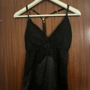 Svart spetslinne från Censored - Supersnygg svart spetslinne från Censored med tunna axelband och spetsdetaljer vid byst och sidor. Linnet har en liten rosett framtill och ett coolt band i ryggen. Materialet är mjukt och glansigt, troligen polyester med spetsinslag. Perfekt för dig som gillar en elegant och edgy look.