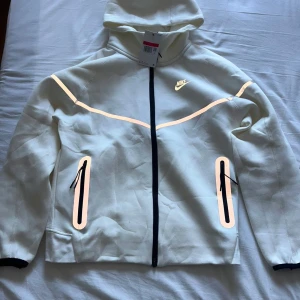 Vit Nike reflective tech fleece  - Nike tracksuit i vitt med matchande hoodie och byxor. Setet har snygga reflexdetaljer i silver och guld längs ärmar och ben, svarta dragkedjor och snörning i midjan. Mjukt material och sportig look med Nike-logga på både jacka och byxa.