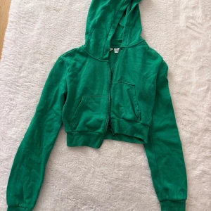 Grön croppad hoodie med dragkedja - Snygg grön croppad hoodie med huva och dragkedja framtill. Tröjan har långa ärmar, ribbade muddar och en stor ficka på magen. Perfekt för dig som gillar en sportig och trendig stil.