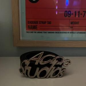 Svart bälte Acne Studios med logospänne - Säljer ett svart bälte från Acne Studios med ett stort, silvrigt logospänne i metall. Bältet är stilrent och tillverkat i slätt skinn, perfekt för att lyfta vilken outfit som helst med en cool detalj. Passar dig som gillar statement-accessoarer. 110 eller 120 cm långt vet inte exakt