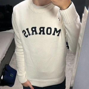 Vit stickad tröja från Morris - Vit stickad tröja från Morris med svart broderad logga på bröstet. Klassisk rund halsringning och ribbade muddar vid ärmslut och nederkant. Snygg detalj med patch på ena ärmen. Perfekt för en clean och stilren look.