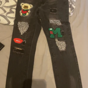 Svarta jeans med patchar och slitningar - Unika svarta jeans med coola patchar som nalle, läppar och bokstaven M. Jeansen har flera slitningar med tyg under och raka ben. Perfekta för dig som vill sticka ut med din stil.