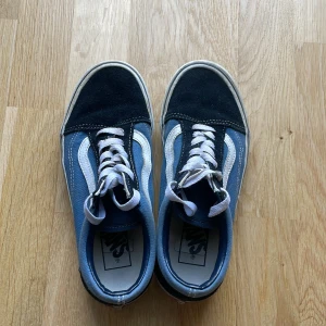 Vans Old Skool blå och svarta sneakers - Klassiska Vans Old Skool sneakers i blått och svart med vit sula och vit sidosnöre. Ovandelen är i canvas och mocka, med vita skosnören och den ikoniska vågiga Vans-loggan på sidan. Perfekta för en avslappnad streetstyle-look.