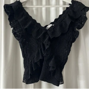 Zara topp - Supersöt svart blus från Zara med djup v-ringning och stora volanger längs halsringningen och axlarna. Blusen är stickad med spetsmönster och har en croppad passform som ger en trendig look. Perfekt att styla med jeans eller kjol för en cool vibe.