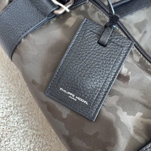 Camouflage resväska från Philippe Model - Snygg resväska från Philippe Model i mörkgrönt camouflage-mönster med svarta skinnhandtag och detaljer. Väskan har dragkedja och en rymlig insida i beige tyg. Perfekt för weekendresor och har en exklusiv känsla med stilrena detaljer.