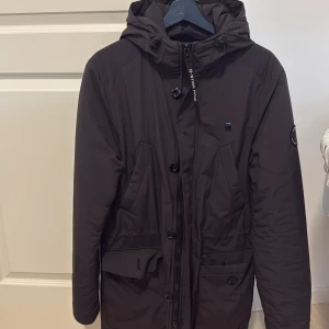 Svart parkajacka från G-Star RAW - Svart parkajacka från G-Star RAW med huva, dragkedja och knappar framtill. Jackan har flera fickor och en diskret logga på bröstet samt ärmen. Perfekt för kalla dagar med sin långa modell och stilrena look.