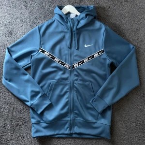 Blå Nike hoodie med dragkedja - Säljer en blå hoodie från Nike med hel dragkedja och huva. Tröjan har svarta och vita band med Nike-logga över bröstet, två fickor framtill och ribbade muddar. Perfekt för dig som gillar sportig och clean stil.