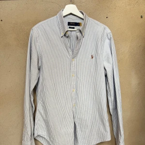 Randig skjorta från Polo Ralph Lauren - Klassisk slim fit skjorta från Polo Ralph Lauren i vitt och blått med tunna vertikala ränder. Skjortan har knappar framtill, lång ärm och broderad logga på bröstet. Tillverkad i bomull och perfekt för en clean och stilren look.
