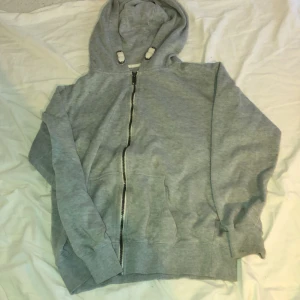 Grå hoodie med dragkedja och huva - Mysig grå hoodie med dragkedja framtill och huva med snörning. Tröjan har långa ärmar, ribbade muddar och en stor ficka på magen. Perfekt för chill dagar eller när du vill vara extra bekväm.