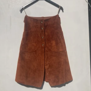 Brun mockakjol från Harders, stl 34 - Snygg brun A-linjeformad kjol i mocka från Harders. Retro 1960-tal. Kjolen har stora fickor fram, knappar längs hela framsidan och bälteshällor. Fodrad insida för extra komfort. Perfekt för dig som gillar retrovibbar och vill sticka ut med en unik look.