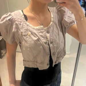  Bolero från Zay - Supersöt ljuslila bolero från Zay med puffärmar, dekorativa knappar och broderade detaljer framtill. Blusen har rundad hals med ljus satinlik kant och små pärlknappar som extra touch.