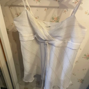 Vit omlott topp från H&M - Supersöt vit topp från H&M med tunna axelband och omlottdetalj framtill. Toppen har subtila glittriga ränder och ett lätt, flowy tyg som ger en luftig känsla. Perfekt för dig som gillar en feminin och stilren look.