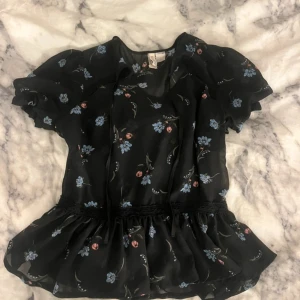 Svart blommig blus från H&M Divided - Söt svart blus från H&M Divided med blå och rosa blommor. Blusen har korta puffärmar, volang nertill och knytband framtill. Oanvänd