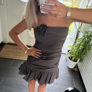 Grå axelbandslös volangklänning - Säljer en grå axelbandslös kortklänning med volangkant nertill och en dekorativ blomdetalj vid bysten. Klänningen har en figurnära passform med rynkade detaljer som ger en snygg siluett. Perfekt för dig som vill sticka ut med en elegant och trendig look.