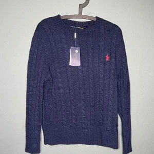 Mörkblå stickad tröja Polo Ralph Lauren - Kabelstickad mörkblå tröja från Polo Ralph Lauren med klassisk rund hals och röd broderad logga på bröstet. Tröjan har lång ärm och ribbade muddar för en snygg passform. Perfekt för dig som gillar stilren och tidlös design.