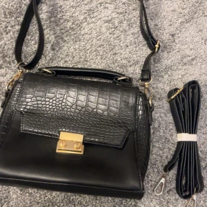 Svart axelväska med krokopräglat lock - Snygg svart axelväska med krokopräglat lock och guldfärgade metalldetaljer. Väskan har både handtag och avtagbar axelrem, samt dragkedja på baksidan. Tillverkad i syntetmaterial med stilren och modern design.