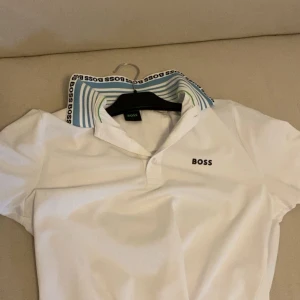 Vit slim fit BOSS t-shirt med krage - Vit kortärmad t-shirt från BOSS i slim fit stretch med klassisk krage. Kragen har blå och gröna ränder samt BOSS-logga runt om. Materialet är mjuk bomull och t-shirten har en stilren BOSS-brodyr på bröstet. Perfekt för dig som gillar clean och sportig stil. Köpte på boozt för 1000. 