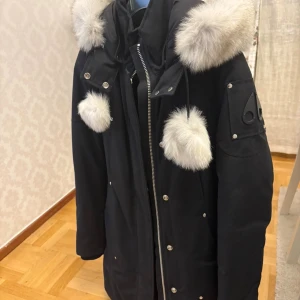 Svart parkajacka med pälsdetaljer Moose Knuckles - Svart parkajacka från Moose Knuckles med silverfärgad dragkedja och tryckknappar. Jackan har stora fluffiga ljusgrå pälsdetaljer på huvan och snörningen. Klassisk logga på ärmen och flera fickor framtill. Perfektl för kalla dagar. 