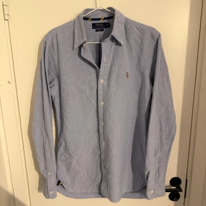 Blå skjorta från Polo Ralph Lauren - Snygg ljusblå skjorta från Polo Ralph Lauren i slim fit-modell. Skjortan har klassisk krage, knäppning framtill och broderad logga på bröstet. Tillverkad i bomull och har långa ärmar. Perfekt för en clean och stilren look.