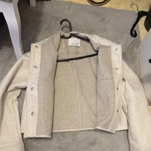 Beige croppad overshirt från Stradivarius - Snygg croppad overshirt i ljusbeige från Stradivarius. Jackan har stora silvriga knappar, klassisk krage och en bröstficka. Tillverkad i mjukt material med ullkänsla och långärmad passform. Perfekt för lager-på-lager och trendig look.