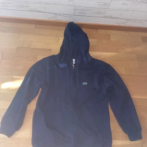Marinblå hoodie från Lacoste L - Säljer en marinblå hoodie från Lacoste i storlek L. Tröjan har dragkedja, huva med snörning och klassisk krokodillogga på bröstet. Tillverkad i 100% bomull och har två fickor framtill. Perfekt för en chill och stilren look.
