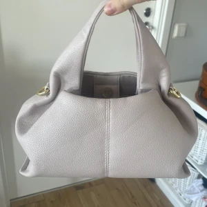 Beige handväska med gulddetaljer - Superfin stilren beige handväska i mjukt material med snygga guldfärgade metalldetaljer. Knappt avvänd i fint skick💕Väskan har en unik, lite puffig form och breda handtag som gör den bekväm att bära. Invändigt finns ett ljust foder och magnetknäppning för enkel stängning. Kan gå ner i pris vid snabb affär!☺️