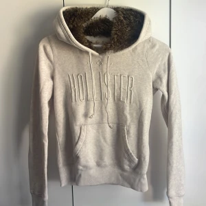 Beige hoodie med fuskpäls från Hollister - Supermysig beige hoodie från Hollister med stor broderad logga framtill. Tröjan har huva fodrad med brun fuskpäls, snörning och klassisk känguruficka. Perfekt för kyliga dagar och riktigt skön att dra på sig. 