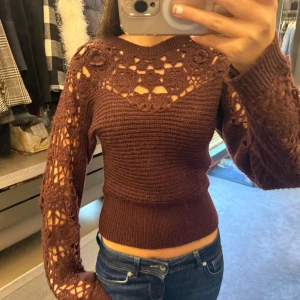 Brun stickad tröja med virkade detaljer - Superfin vinröd/brun stickad tröja med virkade blommor och hålmönster på axlar och ärmar. Tröjan har en tajt midja o ribbad mönster. Bara använt en gång så därför säljs💗