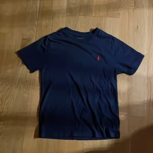 Marinblå t-shirt från Polo Ralph Lauren - Snygg marinblå t-shirt från Polo Ralph Lauren med klassisk röd broderad logga på bröstet. T-shirten är i mjuk bomull och har en normal passform med korta ärmar och rund hals. Perfekt basic-plagg med stilren look.