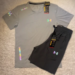 Grå Under Armour set med reflex loggor - Snygg grå Under Armour set med reflex loggor och tryckdetaljer. Det är perfekt för träning. Materialet är lätt och andas, vilket gör den skön att ha på sig under aktiva dagar. Storlek M men passar även S