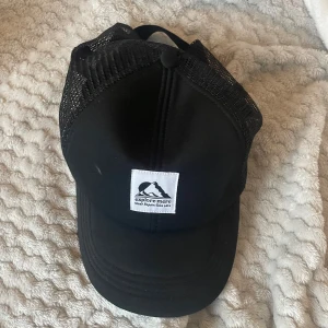 Svart truckerkeps från H&M - Säljer en svart truckerkeps från H&M med vit patch framtill där det står 'explore more' och en bergsillustration. Kepsen har böjd skärm och mesh på sidorna och bak för extra ventilation. Perfekt för dig som gillar enkel och sportig stil.